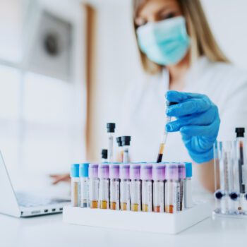 Close up of lab assistant in uniform, with mask and rubber gloves holding test tube with blood sample while sitting on chair and typing on laptop. Selective focus on test tubes.