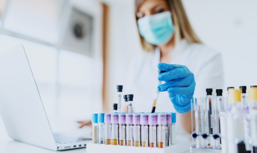 Close up of lab assistant in uniform, with mask and rubber gloves holding test tube with blood sample while sitting on chair and typing on laptop. Selective focus on test tubes.